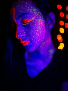 Artistic portrait of a person with neon face paint, illuminated by vibrant colors and bokeh lights.