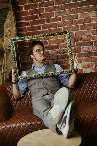 Man sitting on sofa holding picture frame against brick wall backdrop, creative portrait.